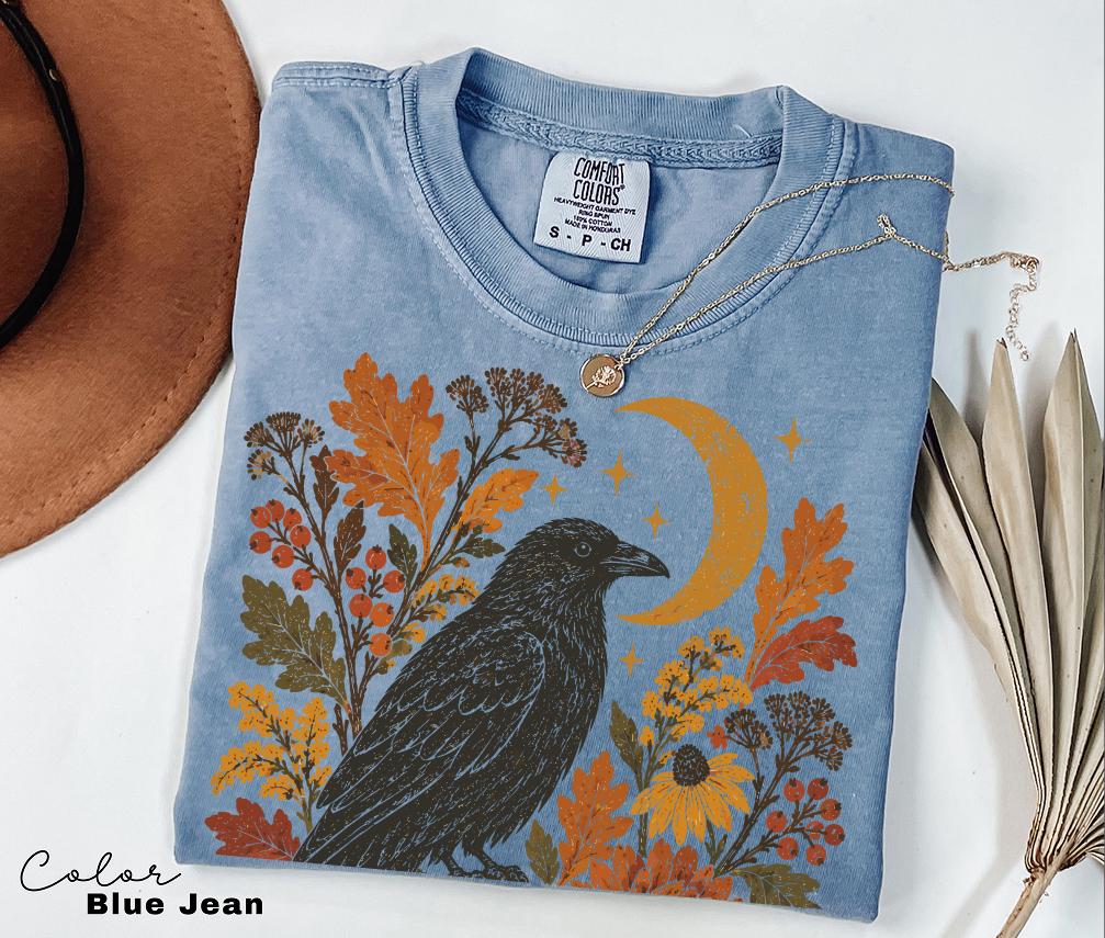 May include: A blue jean colored t-shirt featuring a graphic print of a black crow surrounded by orange autumn leaves, berries, yellow flowers, and a crescent moon. The shirt has a crew neckline and a Comfort Colors brand label at the inner collar.