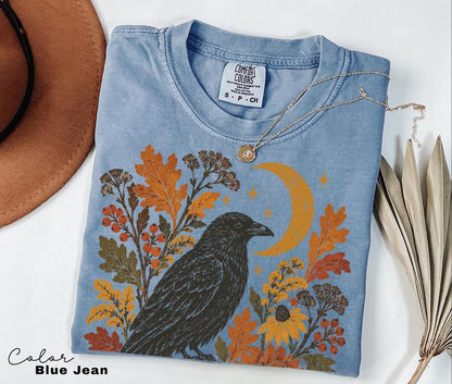 May include: A blue jean colored t-shirt featuring a graphic print of a black crow surrounded by orange autumn leaves, berries, yellow flowers, and a crescent moon. The shirt has a crew neckline and a Comfort Colors brand label at the inner collar.