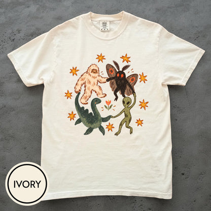 May include: An ivory short-sleeve t-shirt featuring a central graphic of a yeti, a moth-like creature, a green dinosaur, and an alien holding hands in a circle. The design includes orange stars and a small flower, all printed on the front of the shirt.