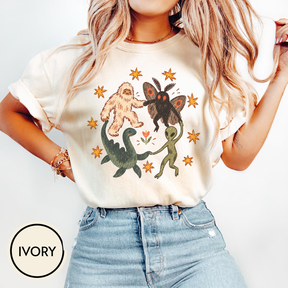 May include: An ivory short-sleeve t-shirt featuring a graphic print of a Bigfoot, a moth-like creature, a green dinosaur, and a green alien holding hands in a circle, surrounded by orange stars and a small flower in the center of the design.