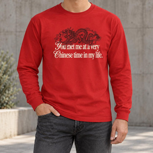 May include: A bright red long-sleeve shirt featuring a black graphic of a dragon positioned above the white text that reads, You met me at a very Chinese time in my life. The shirt has a crew neckline and long sleeves with ribbed cuffs.
