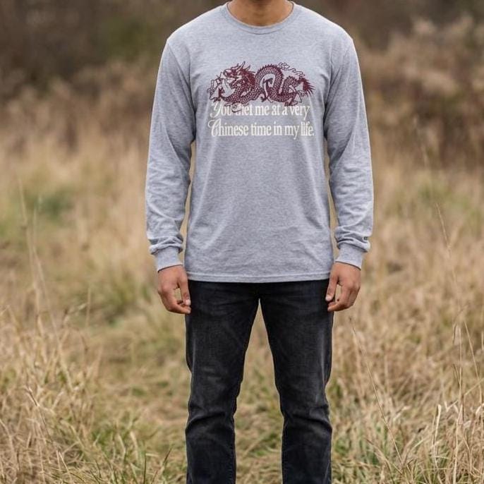 May include: A heather gray long-sleeve shirt featuring a dark red dragon graphic and the white text "You met me at a very Chinese time in my life." The shirt has a crew neckline and long sleeves with ribbed cuffs, paired with dark charcoal denim jeans.