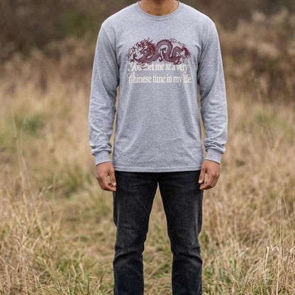May include: A heather gray long-sleeve shirt featuring a dark red dragon graphic and the white text "You met me at a very Chinese time in my life." The shirt has a crew neckline and long sleeves with ribbed cuffs, paired with dark charcoal denim jeans.