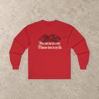 May include: A bright red long-sleeve shirt featuring a black graphic of a dragon positioned above the white text that reads, You met me at a very Chinese time in my life. The shirt has a crew neckline and long sleeves with ribbed cuffs.
