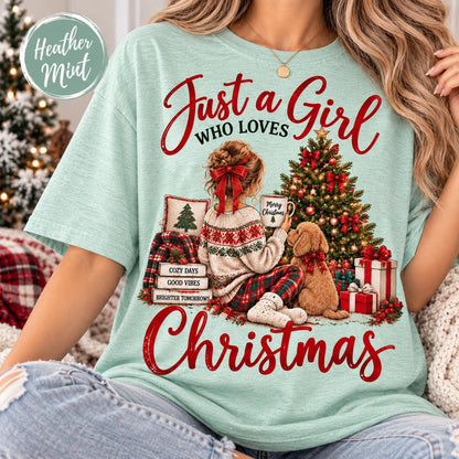 May include: A heather mint t-shirt featuring a graphic of a person sitting by a Christmas tree with a dog. The text reads Just a Girl Who Loves Christmas, Merry Christmas, Cozy Days, Good Vibes, and Brighter Tomorrows. The design includes gifts.