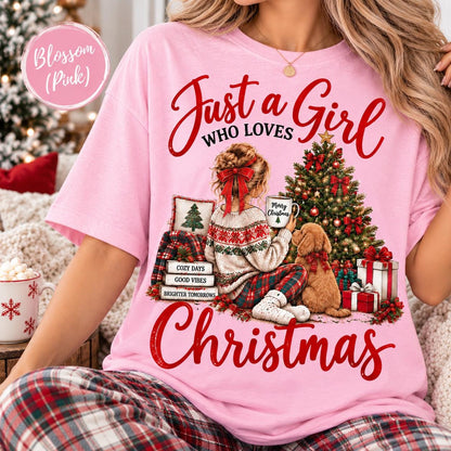 May include: A light pink t-shirt featuring a graphic of a person with a red hair bow sitting by a Christmas tree with a dog. The text reads Just a Girl WHO LOVES CHRISTMAS, Merry Christmas, COZY DAYS, GOOD VIBES, and BRIGHTER TOMORROWS.