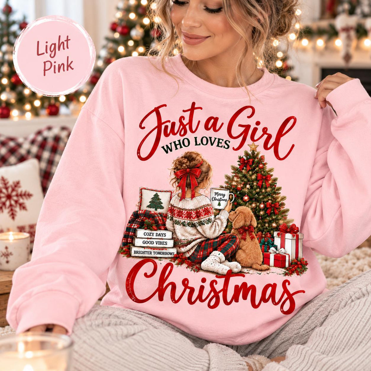 May include: A light pink sweatshirt featuring a graphic of a person with a red hair bow sitting by a Christmas tree with a dog. The text reads Just a Girl WHO LOVES Christmas, with stacked books labeled COZY DAYS, GOOD VIBES, BRIGHTER TOMORROWS.