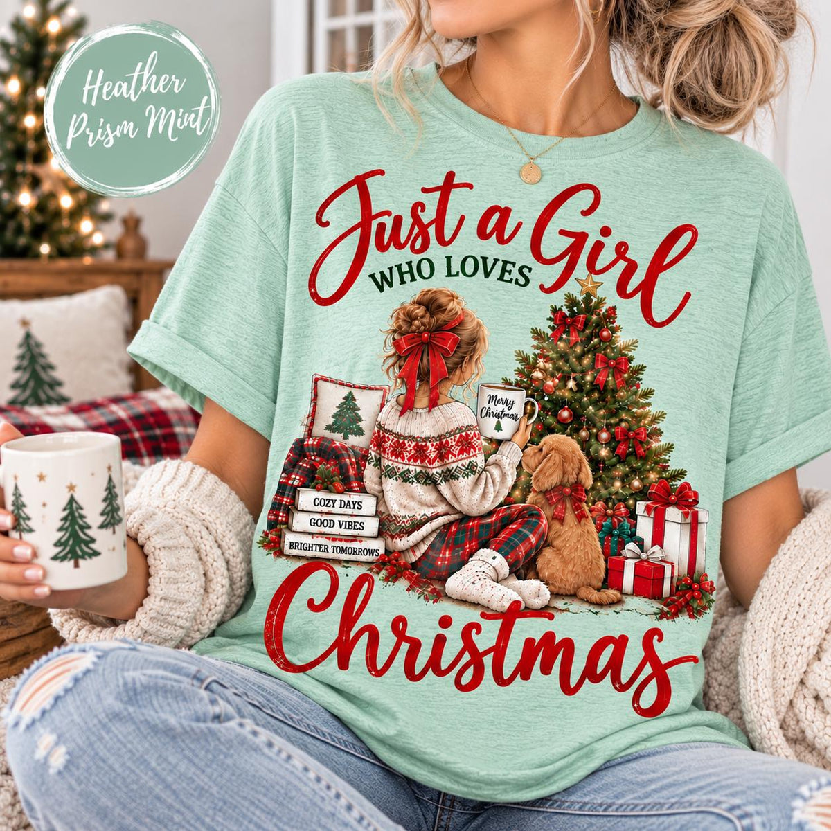 May include: A heather prism mint t-shirt featuring a graphic of a person with a red bow in their hair sitting by a Christmas tree with a dog. The text reads Just a Girl WHO LOVES CHRISTMAS, with smaller text on stacked pillows reading COZY DAYS, GOOD VIBES, BRIGHTER TOMORROWS.