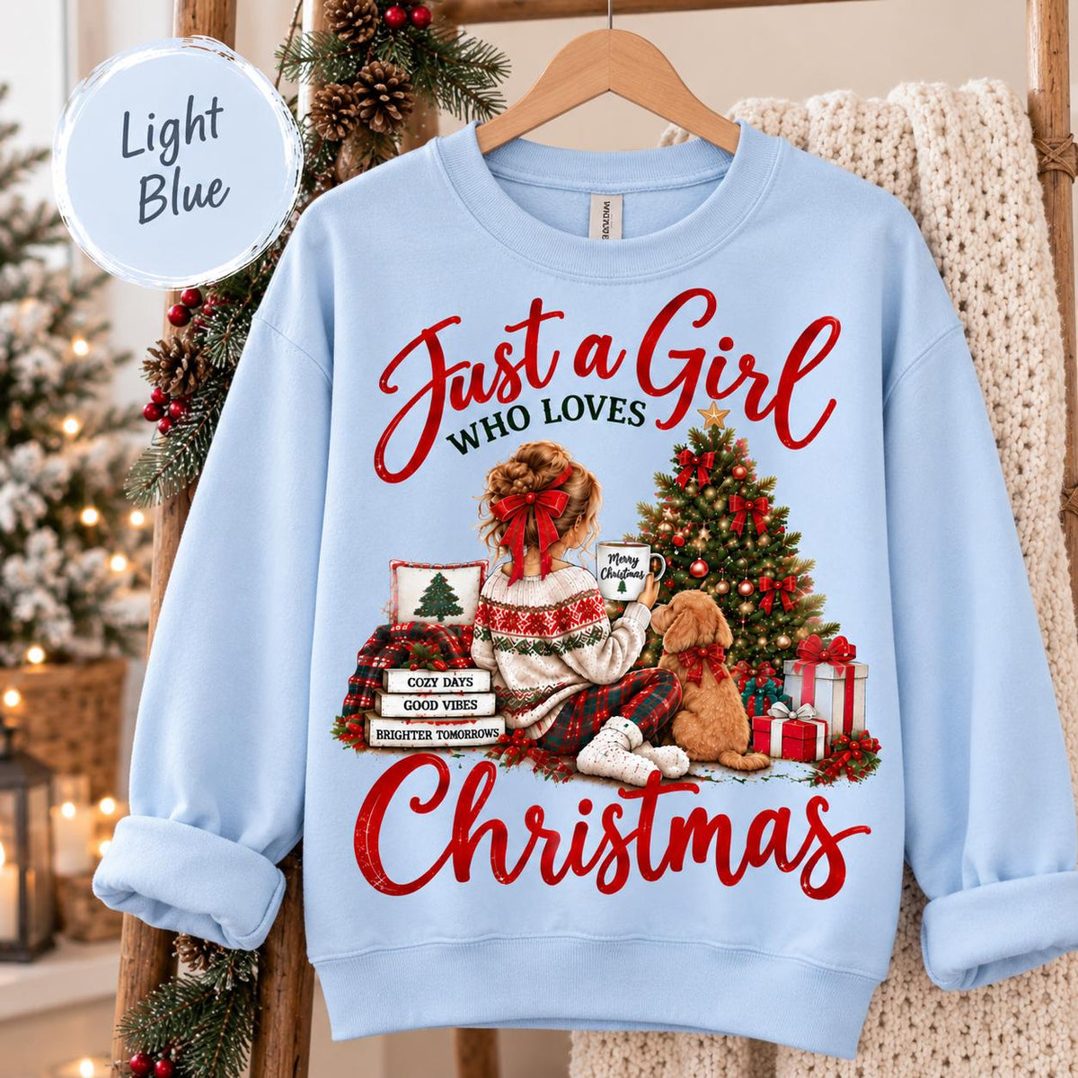 May include: A light blue crewneck sweatshirt featuring a graphic print of a person with a red hair bow sitting next to a dog and a Christmas tree. The text reads Just a Girl WHO LOVES Christmas, with smaller text on stacked books reading COZY DAYS, GOOD VIBES, BRIGHTER TOMORROWS.