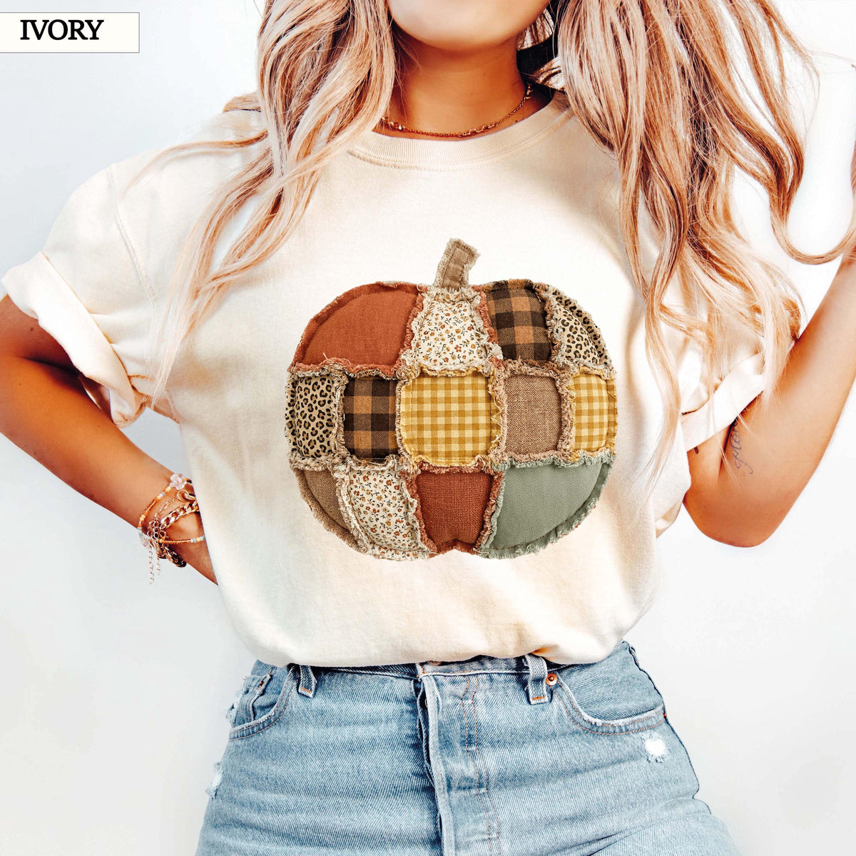 May include: An ivory t-shirt featuring a central graphic of a pumpkin constructed from various fabric patches. The pumpkin design includes brown, tan, and green squares with patterns such as leopard print, buffalo plaid, and gingham checks.