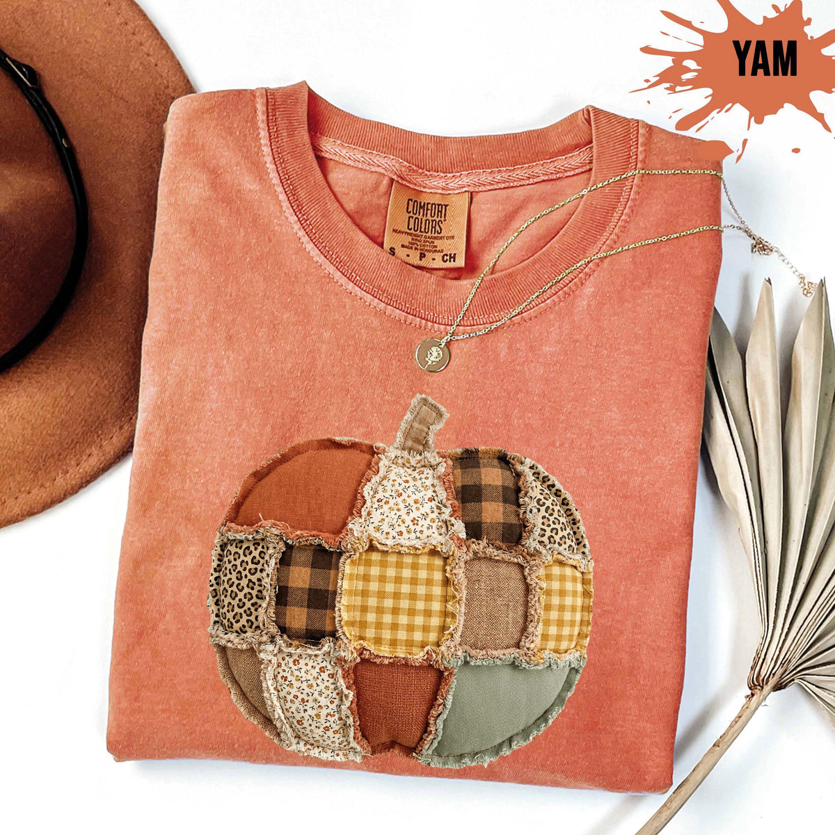 May include: A terracotta-colored t-shirt featuring a central pumpkin graphic composed of various fabric patches, including plaid, leopard print, and floral patterns. The shirt has a crew neckline and a Comfort Colors label at the inner collar.