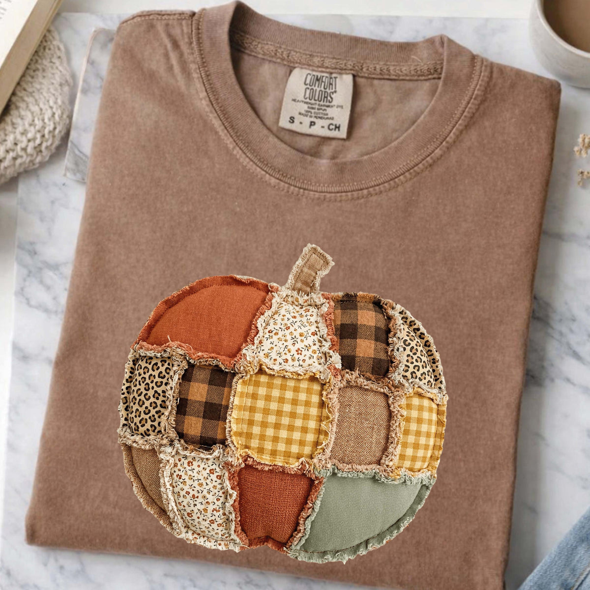May include: A brown crew neck t-shirt featuring a central pumpkin graphic made of various fabric patches. The pumpkin design includes squares of orange, yellow gingham, brown plaid, leopard print, and floral patterns with frayed edges.
