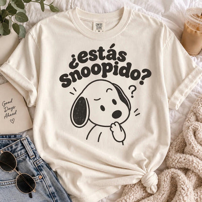 May include: A cream-colored short-sleeve t-shirt featuring a black graphic of a cartoon dog with one ear down and a hand to its chin. Above the dog, the text reads ¿estás Snoopido? in a bold, rounded font. The shirt has a crew neckline.
