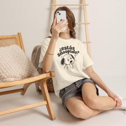 May include: A cream-colored short-sleeve t-shirt featuring a black graphic of a cartoon dog with the text "¿estás snoopido?" printed above it. The shirt has a crew neckline and a relaxed fit, paired with dark gray denim shorts.