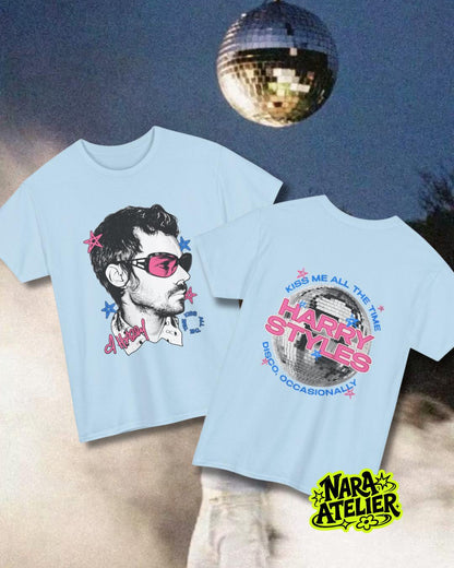 May include: A light blue t-shirt featuring a graphic of a person wearing pink sunglasses on the front and a disco ball graphic with the text "HARRY STYLES KISS ME ALL THE TIME DISCO, OCCASIONALLY" on the back, accented with small pink and blue stars.