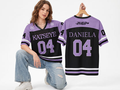 May include: A black and lavender jersey featuring the text KATSEYE and the number 04 on the front, with the text WILD, DANIELA, and the number 04 on the back. The jersey has short sleeves with white and black stripes and a V-neck collar.