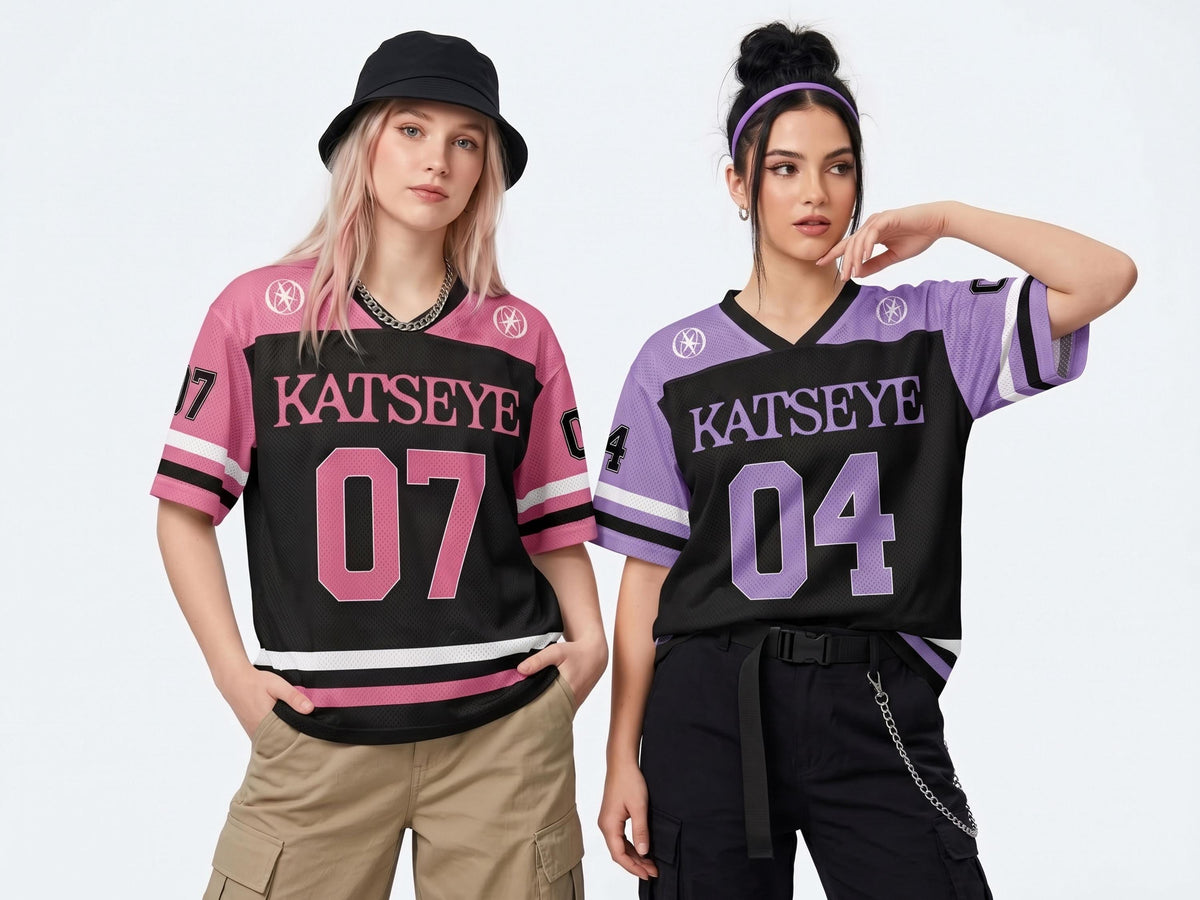 May include: Two short-sleeve jerseys featuring the text KATSEYE. One jersey is pink and black with the number 07, while the other is purple and black with the number 04. Both shirts have V-necks, striped sleeve cuffs, and circular logos on the shoulders.