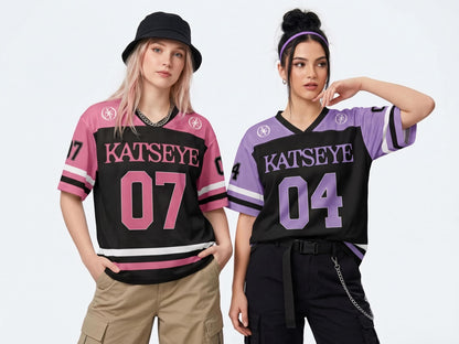 May include: Two short-sleeve jerseys featuring the text KATSEYE. One jersey is pink and black with the number 07, while the other is purple and black with the number 04. Both shirts have V-necks, striped sleeve cuffs, and circular logos on the shoulders.