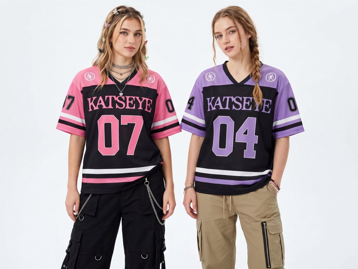 May include: Two V-neck jerseys featuring the text KATSEYE across the chest. One jersey is pink and black with the number 07, while the other is purple and black with the number 04. Both shirts have short sleeves, striped cuffs, and circular logos.