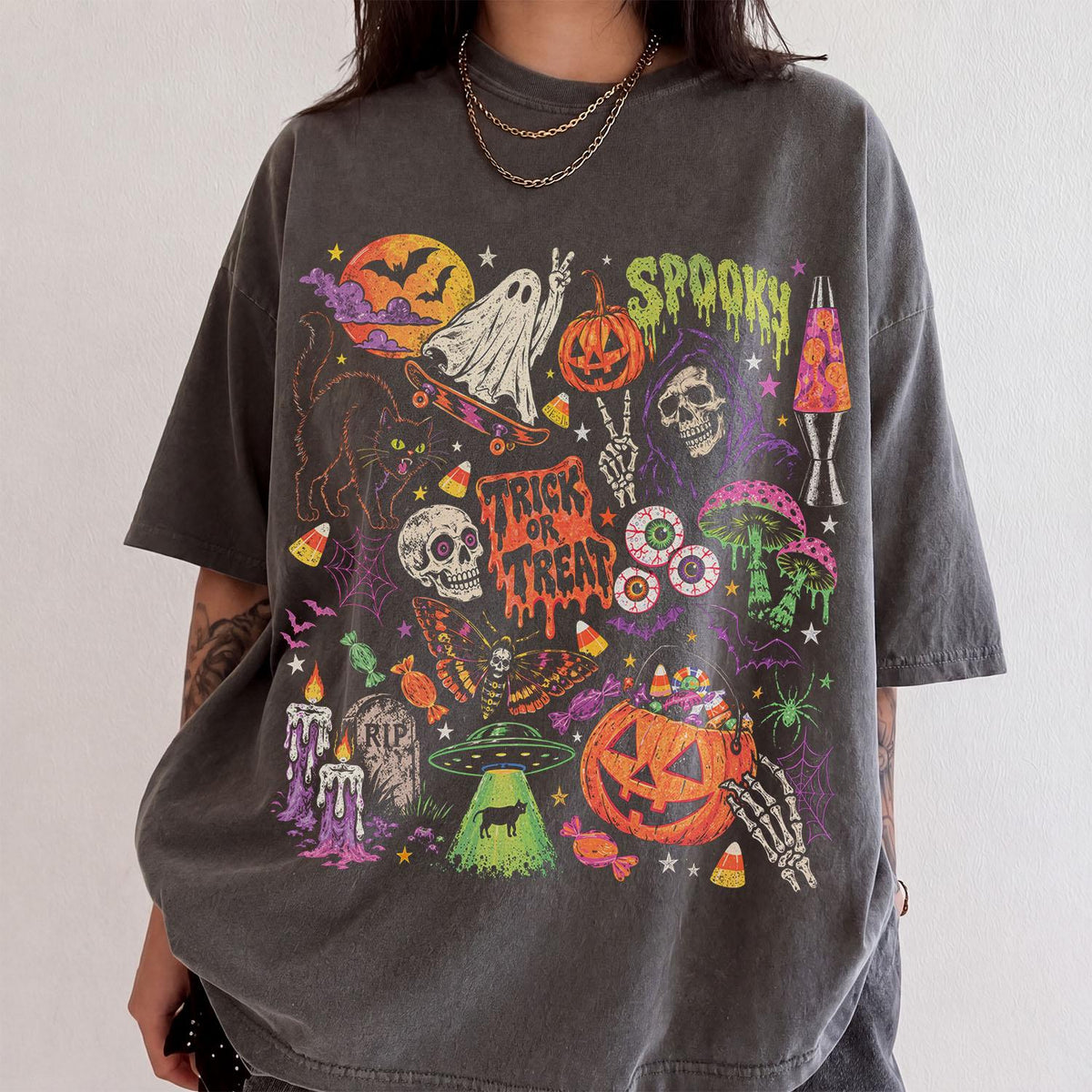 May include: A charcoal gray t-shirt featuring a vibrant Halloween-themed graphic print. The design includes a ghost on a skateboard, a skull, a jack-o'-lantern, a black cat, a moth, a UFO, melting text reading TRICK OR TREAT, and SPOOKY.