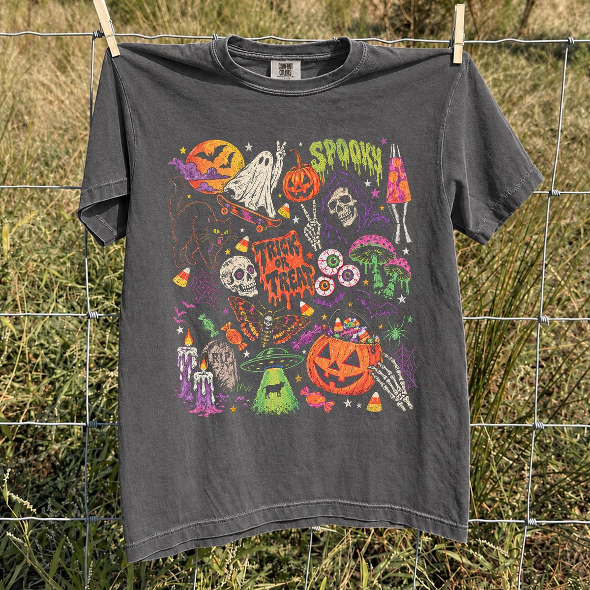 May include: A charcoal gray short-sleeve t-shirt featuring a vibrant, multi-colored Halloween-themed graphic print. The design includes a ghost on a skateboard, a pumpkin, a skull, a UFO, candy corn, and the text "TRICK OR TREAT" and "SPOOKY".