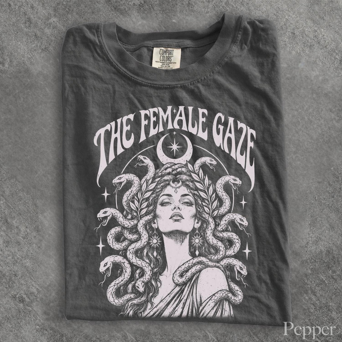 May include: A charcoal gray t-shirt featuring a white graphic print of a person with snakes for hair, reminiscent of Medusa. Above the figure, the text reads THE FEMALE GAZE in a curved font. A crescent moon and stars surround the central illustration.
