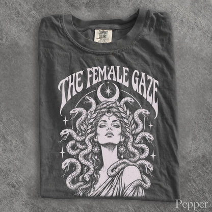 May include: A charcoal gray t-shirt featuring a white graphic print of a person with snakes for hair, reminiscent of Medusa. Above the figure, the text reads THE FEMALE GAZE in a curved font. A crescent moon and stars surround the central illustration.