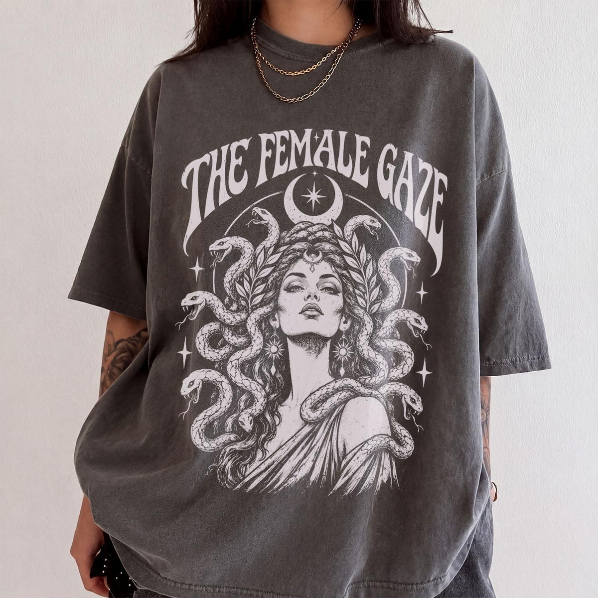 May include: A charcoal gray short-sleeve t-shirt featuring a white graphic print of a figure with snakes for hair. The text "THE FEMALE GAZE" is printed in an arched font above the figure, accompanied by a crescent moon and star symbols.