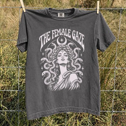 May include: A charcoal gray short-sleeve t-shirt featuring a white graphic print of a person with snakes for hair, surrounded by a crescent moon and stars. The text THE FEMALE GAZE is arched above the illustration in a decorative serif font.