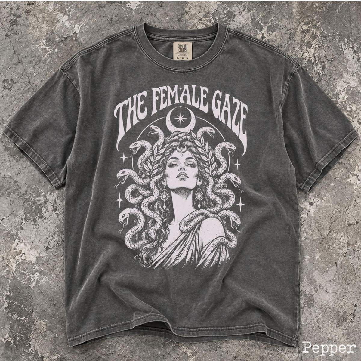 May include: A charcoal gray short-sleeve t-shirt featuring a white graphic print of a person with snakes for hair, surrounded by stars and a crescent moon. The text THE FEMALE GAZE is arched above the illustration in a bold, serif font.