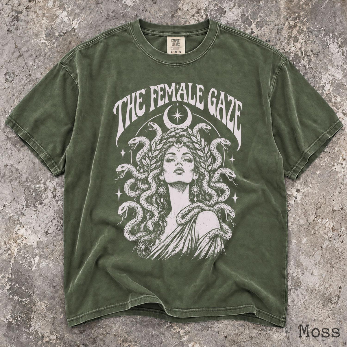 May include: A moss green short-sleeve t-shirt featuring a white graphic print of a person with snakes for hair. The text THE FEMALE GAZE is arched above the figure, which is framed by a crescent moon and stars. The shirt has a crew neckline.