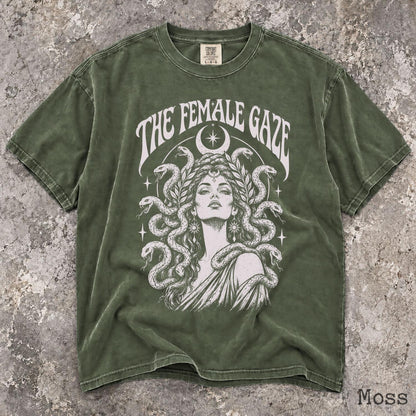 May include: A moss green short-sleeve t-shirt featuring a white graphic print of a person with snakes for hair. The text THE FEMALE GAZE is arched above the figure, which is framed by a crescent moon and stars. The shirt has a crew neckline.
