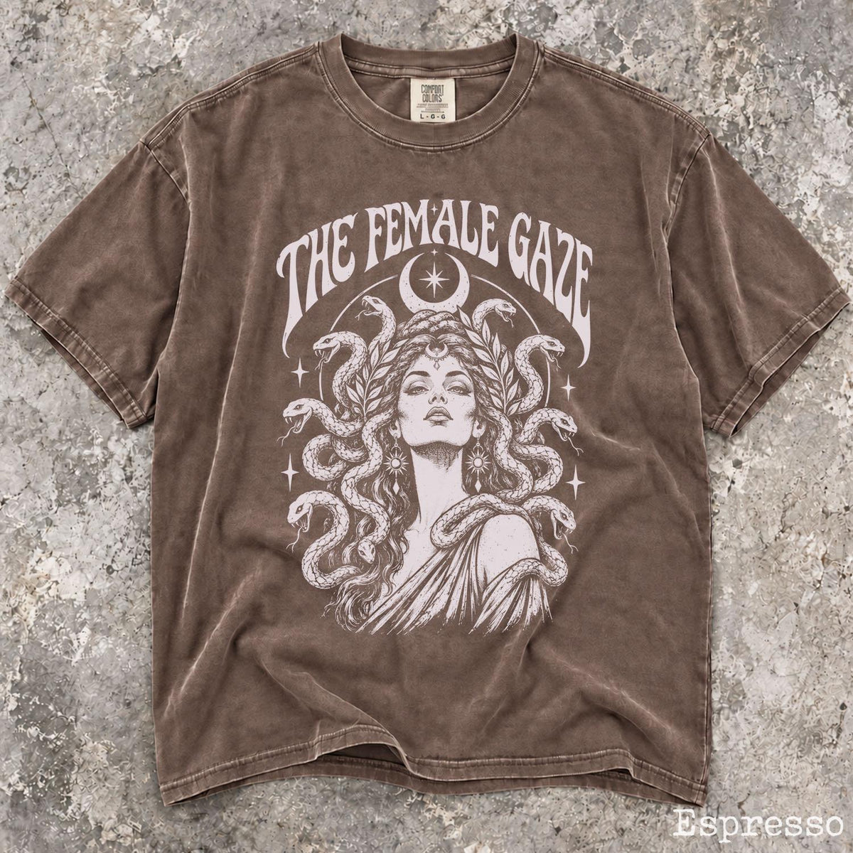 May include: A brown short-sleeve t-shirt featuring a graphic print of a person with snakes for hair, a crescent moon, and stars. The text THE FEMALE GAZE is printed in white above the illustration. The fabric has a washed, vintage-style finish.