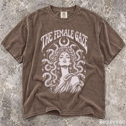 May include: A brown short-sleeve t-shirt featuring a graphic print of a person with snakes for hair, a crescent moon, and stars. The text THE FEMALE GAZE is printed in white above the illustration. The fabric has a washed, vintage-style finish.