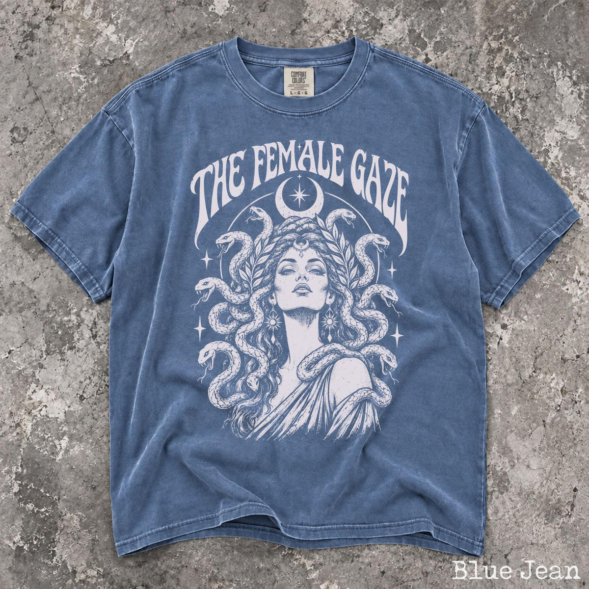 May include: A blue jean colored t-shirt featuring a white graphic print of a person with snakes for hair, surrounded by stars and a crescent moon. The text THE FEMALE GAZE is printed in a curved font above the illustration on the front of the shirt.