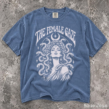 May include: A blue jean colored t-shirt featuring a white graphic print of a person with snakes for hair, surrounded by stars and a crescent moon. The text THE FEMALE GAZE is printed in a curved font above the illustration on the front of the shirt.