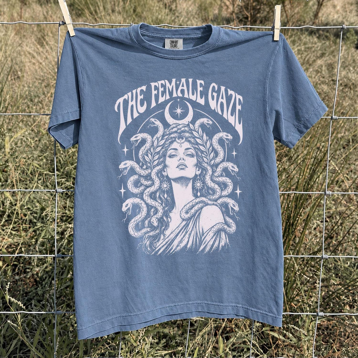 May include: A blue short-sleeve t-shirt featuring a white graphic print of a person with snakes for hair, surrounded by stars and a crescent moon. The text THE FEMALE GAZE is printed in a curved arch above the central figure on the front of the shirt.