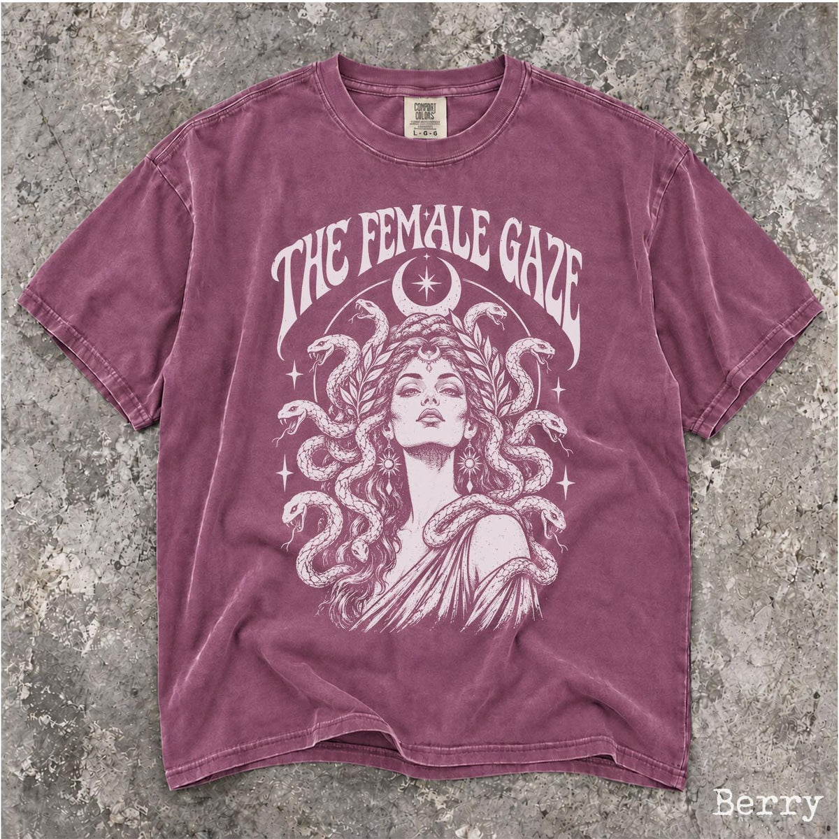May include: A berry-colored short-sleeve t-shirt featuring a white graphic print of a figure with snakes for hair. The text THE FEMALE GAZE is arched above the figure, with a crescent moon and stars surrounding the design. The shirt has a crew neck.