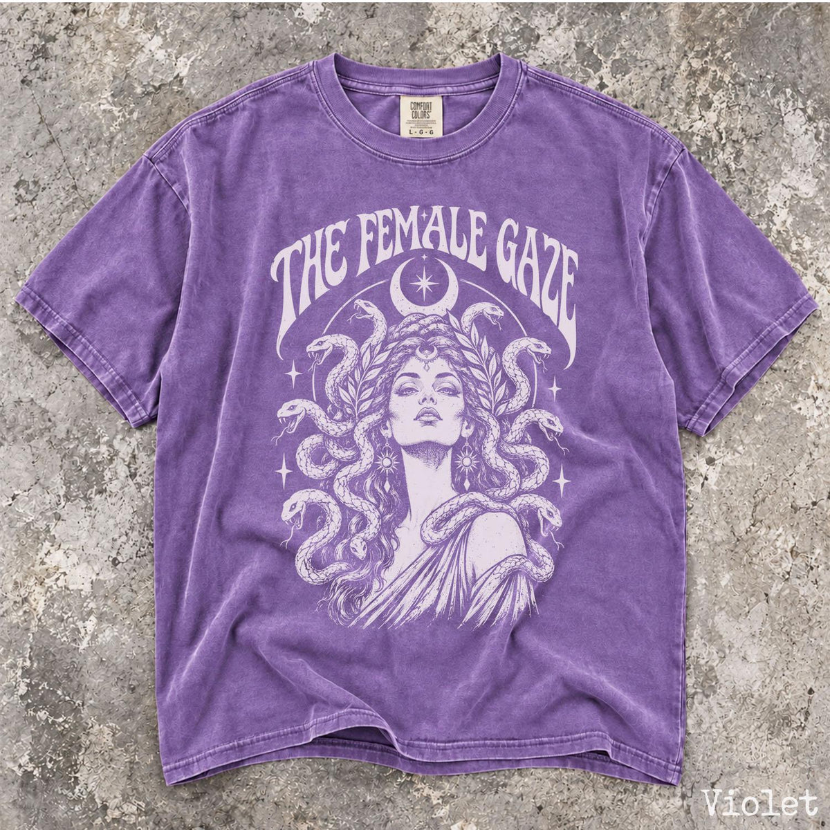May include: A violet short-sleeve t-shirt featuring a white graphic print of a person with snakes for hair, surrounded by stars and a crescent moon. The text THE FEMALE GAZE is printed in an arched font above the illustration on the front.