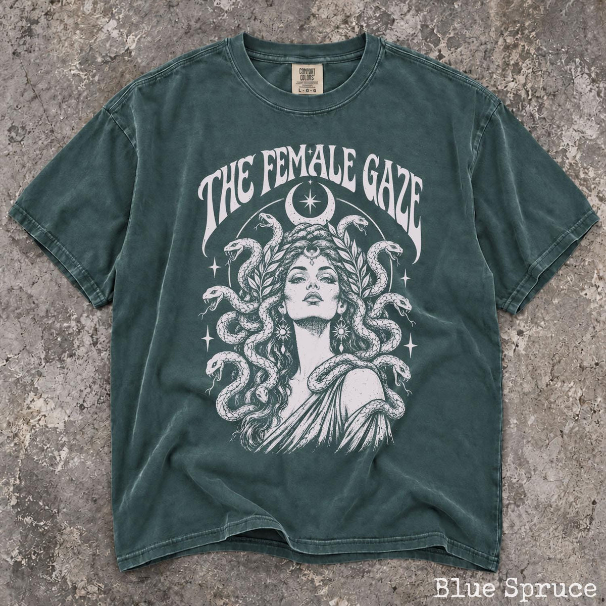 May include: A blue spruce t-shirt featuring a white graphic print of a person with snakes for hair, a crescent moon, and stars. The text THE FEMALE GAZE is printed in an arched font above the illustration. The shirt has a crew neck and short sleeves.