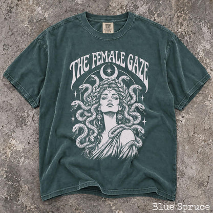 May include: A blue spruce t-shirt featuring a white graphic print of a person with snakes for hair, a crescent moon, and stars. The text THE FEMALE GAZE is printed in an arched font above the illustration. The shirt has a crew neck and short sleeves.