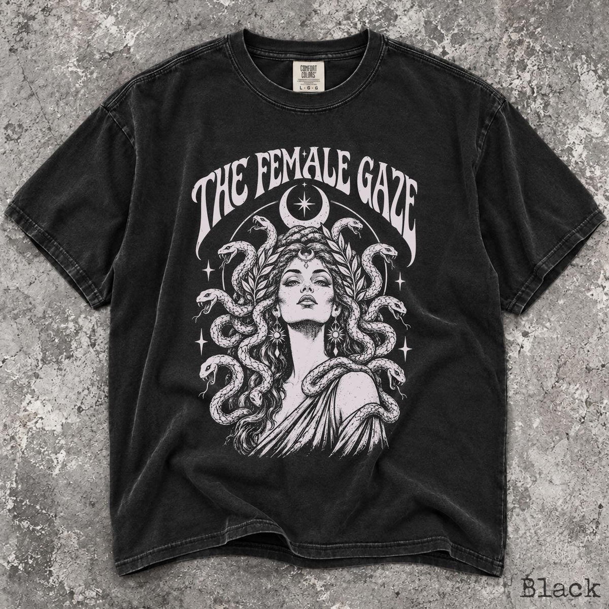 May include: A black short-sleeve t-shirt featuring a white graphic print of a person with snakes for hair, surrounded by stars and a crescent moon. The text THE FEMALE GAZE is arched above the illustration. The shirt has a crew neckline and a faded finish.
