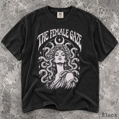 May include: A black short-sleeve t-shirt featuring a white graphic print of a person with snakes for hair, surrounded by stars and a crescent moon. The text THE FEMALE GAZE is arched above the illustration. The shirt has a crew neckline and a faded finish.