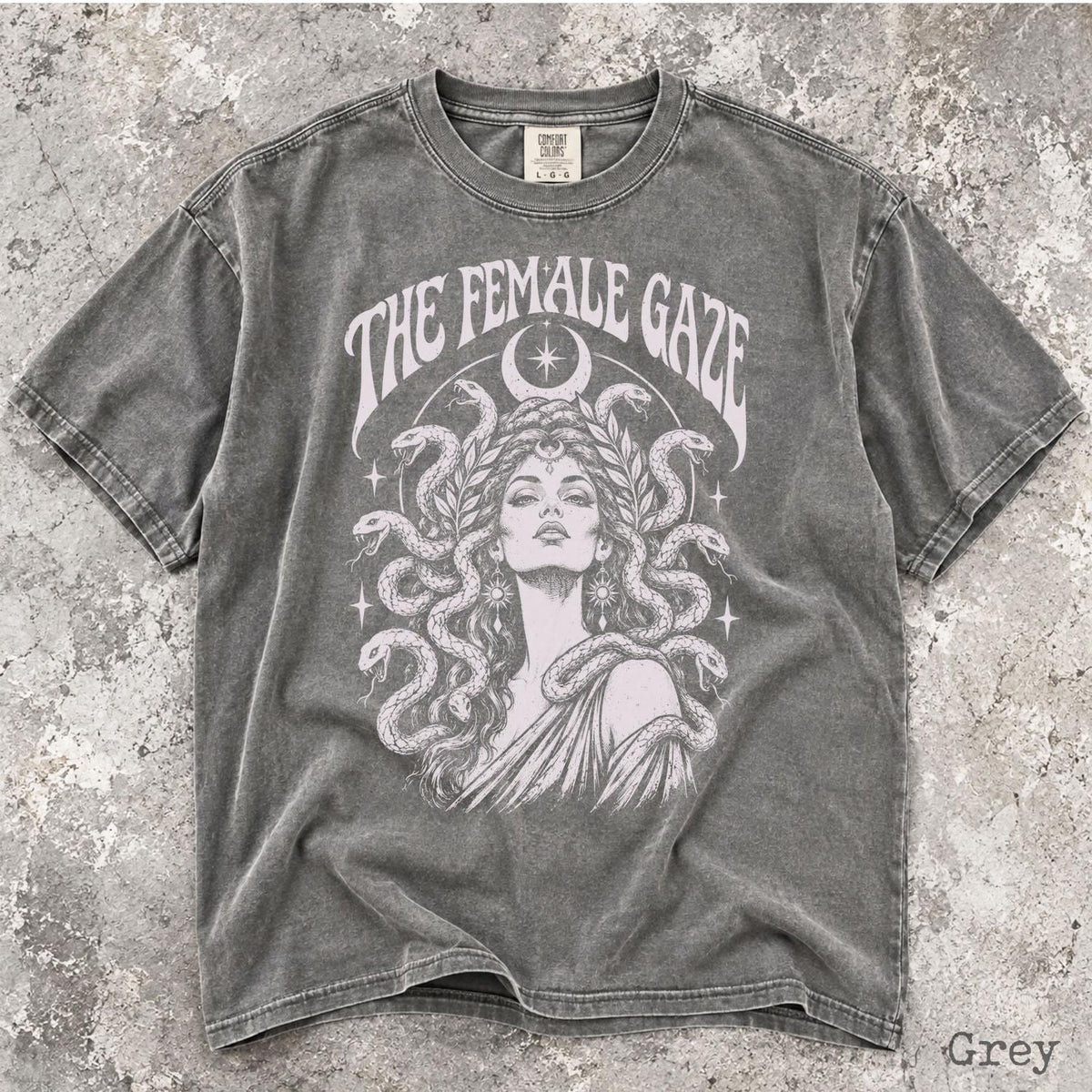 May include: A charcoal grey short-sleeve t-shirt featuring a graphic print of a person with snakes for hair, surrounded by stars and a crescent moon. The text THE FEMALE GAZE is printed in an arched font above the illustration on the front.