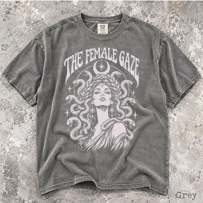 May include: A charcoal grey short-sleeve t-shirt featuring a graphic print of a person with snakes for hair, surrounded by stars and a crescent moon. The text THE FEMALE GAZE is printed in an arched font above the illustration on the front.