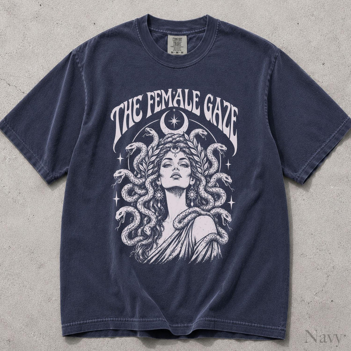 May include: A navy blue short-sleeve t-shirt featuring a white graphic print of a person with snakes for hair, surrounded by laurel leaves and a crescent moon. The text THE FEMALE GAZE is printed in an arch above the central illustration.