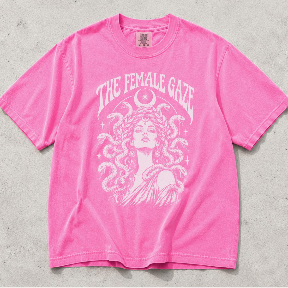 May include: A bright pink short-sleeve t-shirt featuring a white graphic print of a person with snakes for hair, surrounded by stars and a crescent moon. The text THE FEMALE GAZE is printed in a curved font above the central illustration.