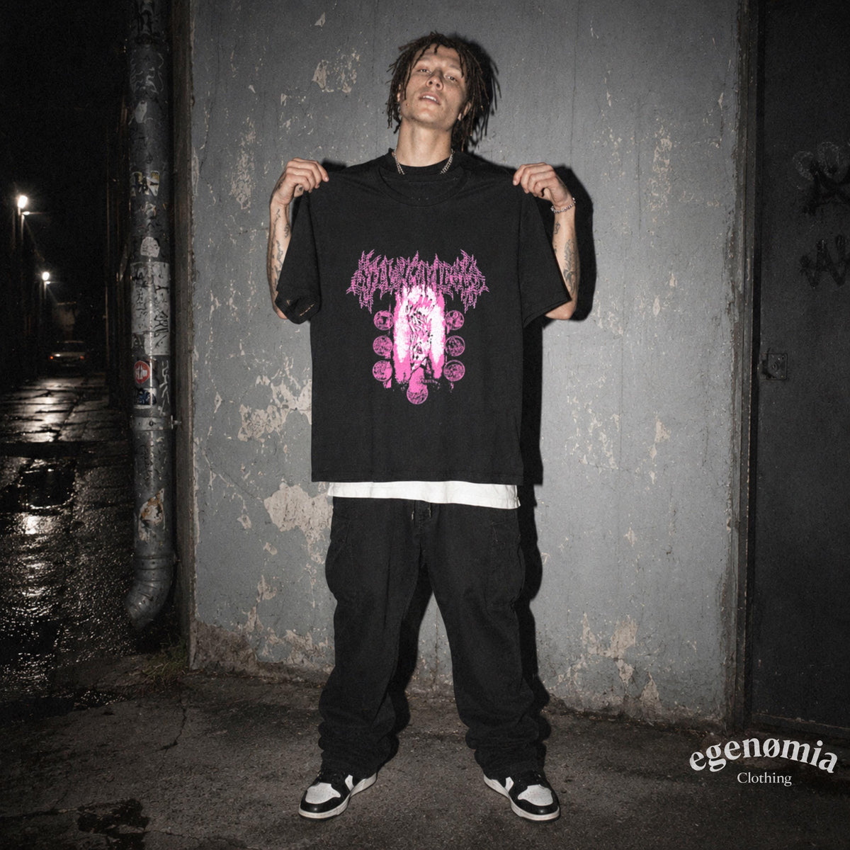 May include: A black short-sleeve t-shirt featuring a pink graphic print on the chest with stylized text and circular motifs. The shirt is worn over a white undershirt, paired with loose-fitting black pants and black and white sneakers.