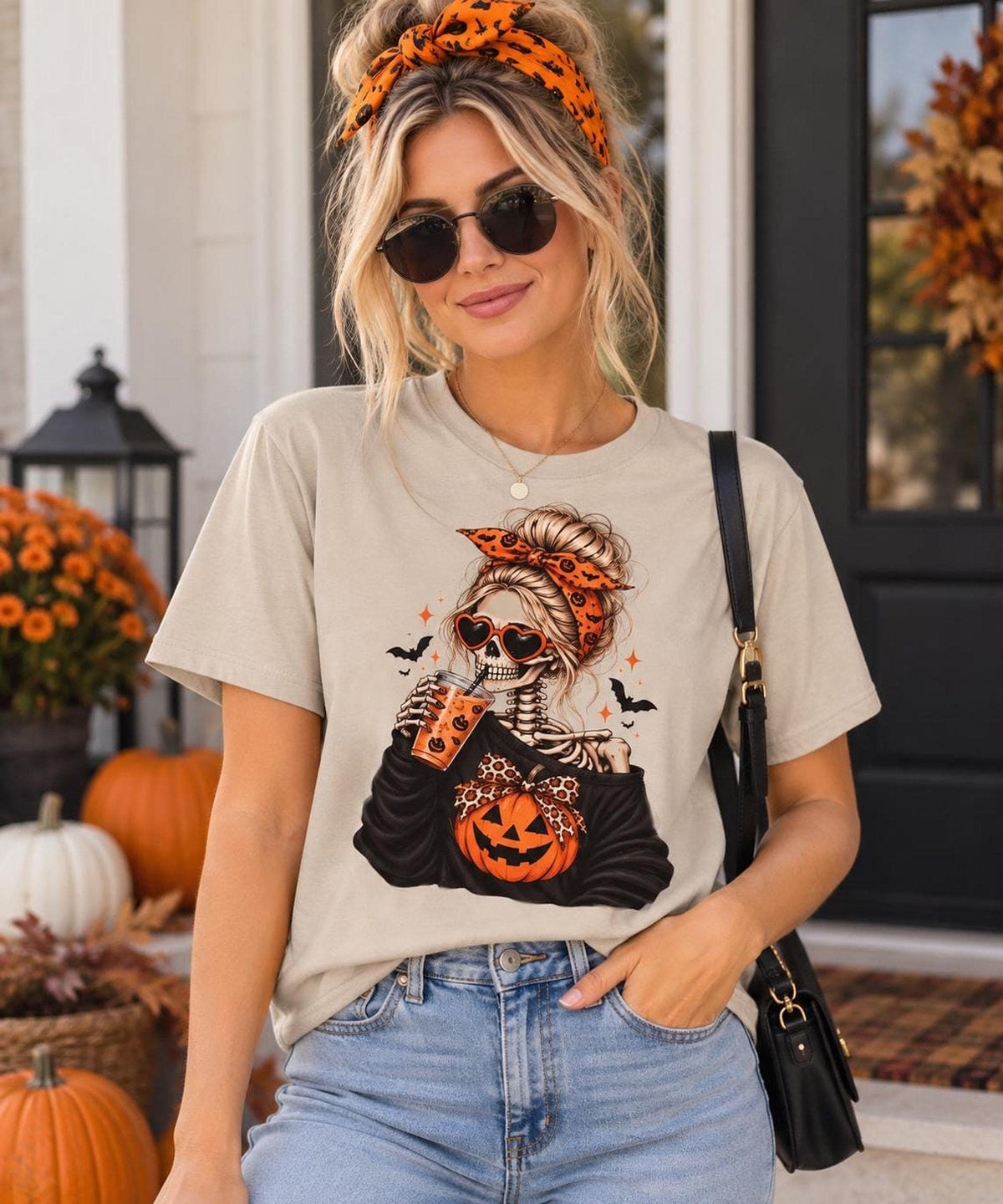 A blonde woman wearing sunglasses, an orange headband, and a beige t-shirt featuring a female skeleton graphic holding an iced drink.