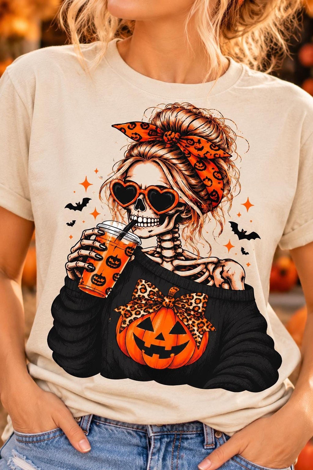 A woman wearing a sand-colored t-shirt featuring a graphic of a skeletal woman with heart sunglasses and a pumpkin.